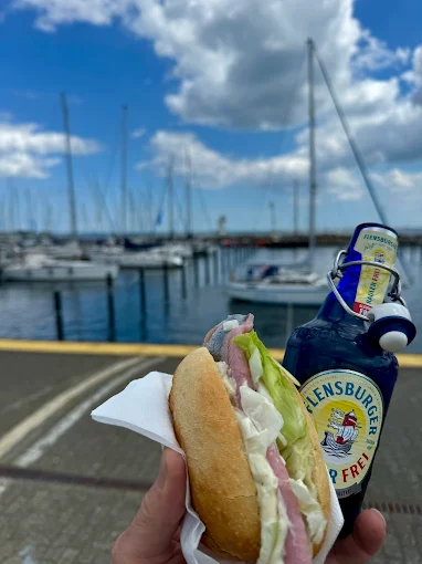 Ein leckeres Fischbrötchen in der Hand mit dem Hafen von Schilksee im Hintergrund